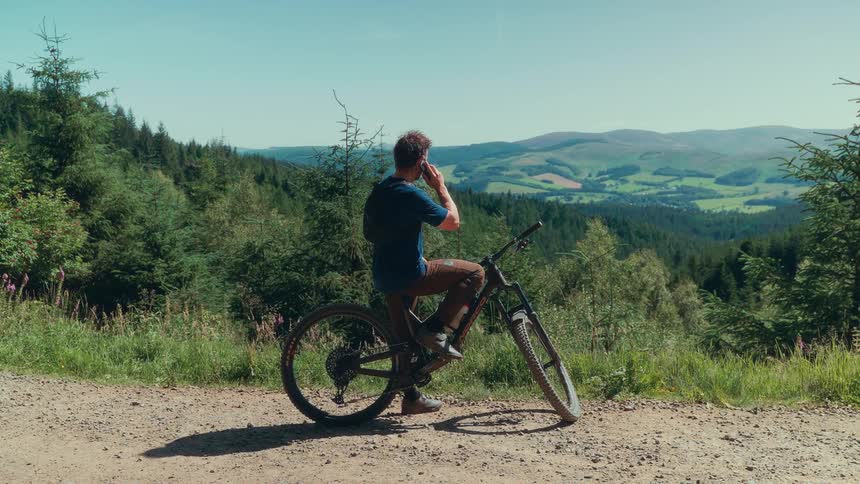 A man talks on the phone while seated on a mountain bike on a dirt path overlooking rolling green hills and forested valleys under a clear sky