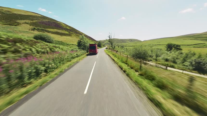 A red van drives along a narrow country road surrounded by green hills and fields under a bright sky