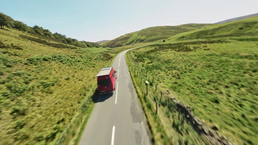 Red van drives along narrow countryside road flanked by green fields and distant hills, under a bright sunny sky