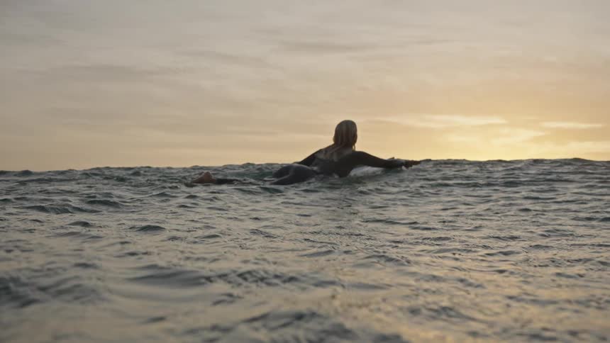 A surfer paddles on the ocean at sunset