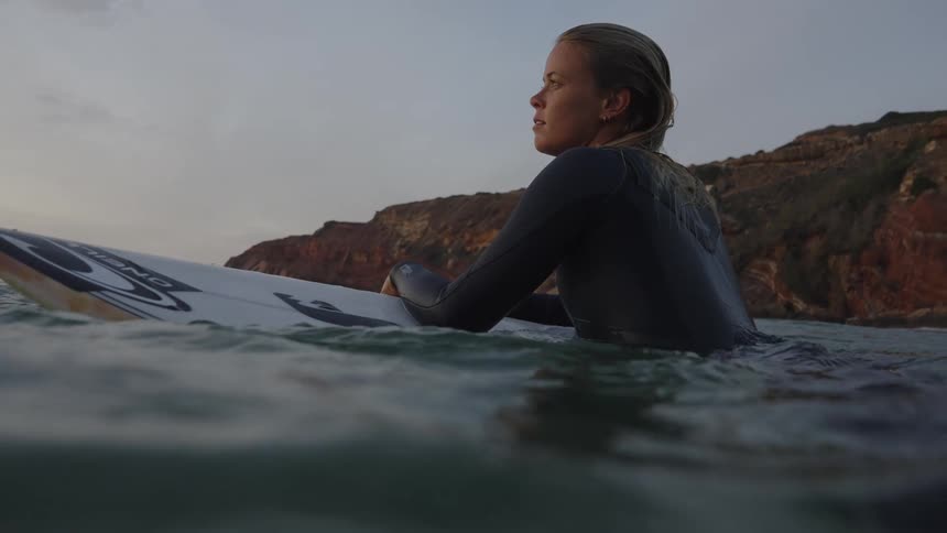 A surfer rests on her board in the ocean by cliffs at sunset