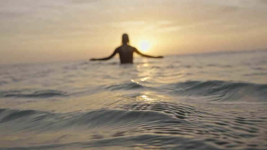A woman stands in the ocean submerged from the waist down as waves crash around her at sunset