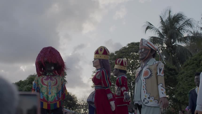 The video shows a parade or outdoor gathering featuring several large humanlike statues or effigies dressed in colorful costumes the figures are distinct with one wearing a red dress and a crown and others in traditional or ornate outfits the scene