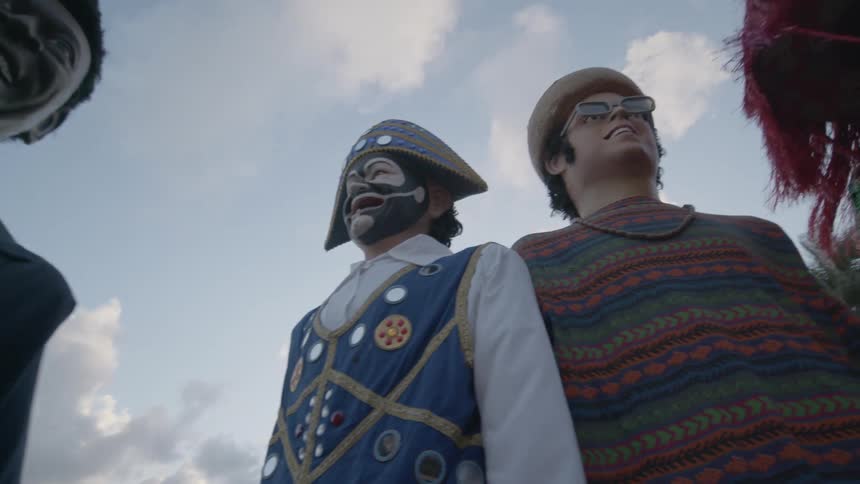 A scene outside a castle with people wearing costumes with big masks dancing as the camera is in the middle of the circle