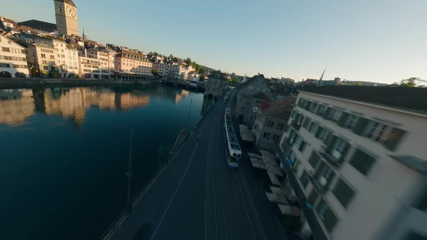 An aerial view of a european city shows a river flowing through it with buildings lining both banks the shot follows a modern tram as it moves along a bridge or street beside the water with a prominent clock tower visible on the far bank