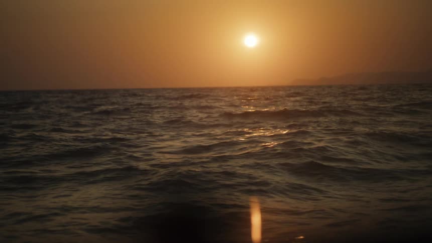 The orange sun sets over a calm rippling ocean with distant mountains faintly visible