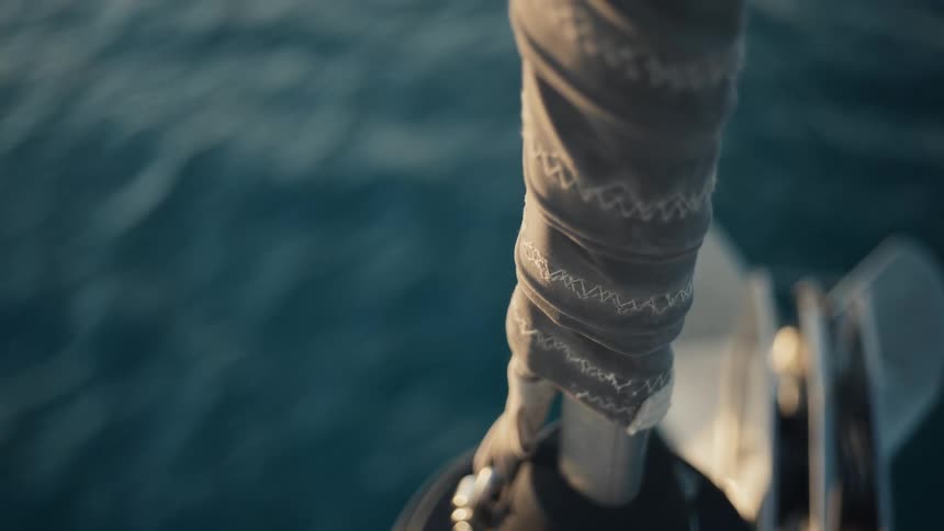 The video shows a highangle shot of a deep blue body of water with small waves on the surface in the foreground out of focus a person is holding onto a rope or a part of a boat with what appears to be a lightcolored patterned fabric wrapped around