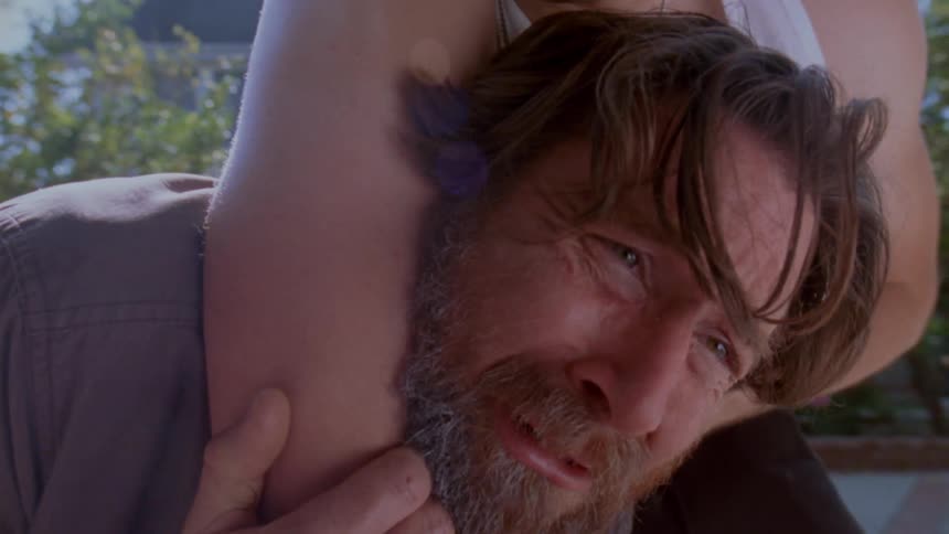 A man with a beard and brown hair looks up while being held in a headlock then a house is revealed