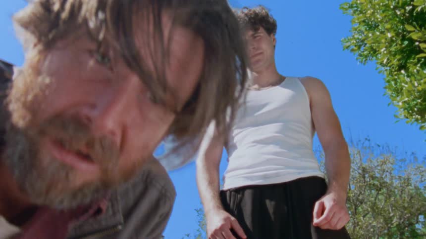 The face of a bearded man who has an expression of suffering and he is crouched on the ground and behind him there is a man in a white tank top watching