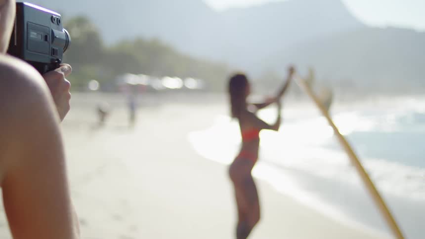 The video shows a person holding a vintage film camera filming a woman on a sunny beach the woman being filmed who is in the background is wearing an orange bikini has tattoos on her side and legs and is holding a long yellow surfboard