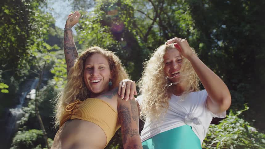 Two smiling women with long curly blonde hair standing close together in a lush green outdoor setting