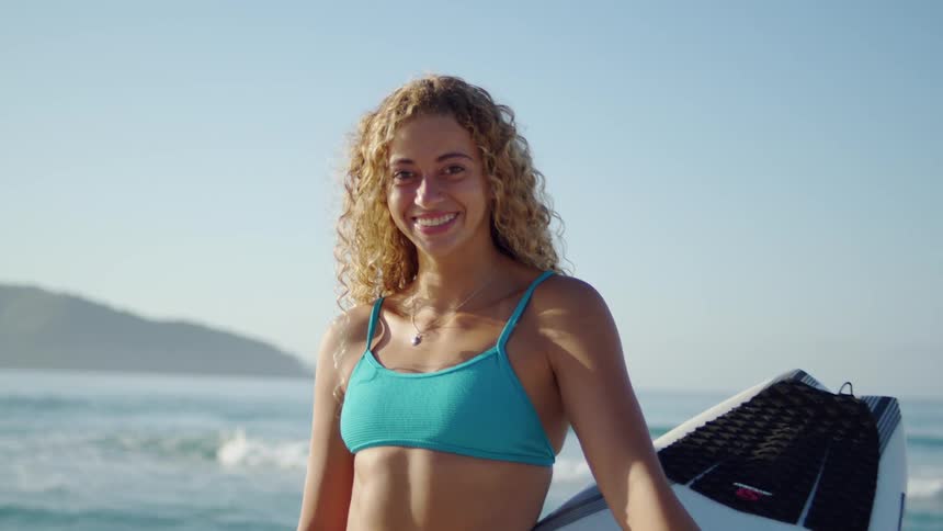 A medium shot shows a young woman with curly blonde hair smiling directly at the camera she is wearing a blue bikini top and is holding a surfboard next to her on a sunny beach