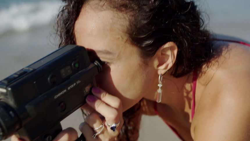 A person is holding and looking through a handheld video camera on a beach the woman is wearing a red top and silver hoop earrings
