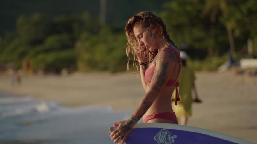 A young woman wearing a bikini stands on the beach holding a surfboard on a sunny day