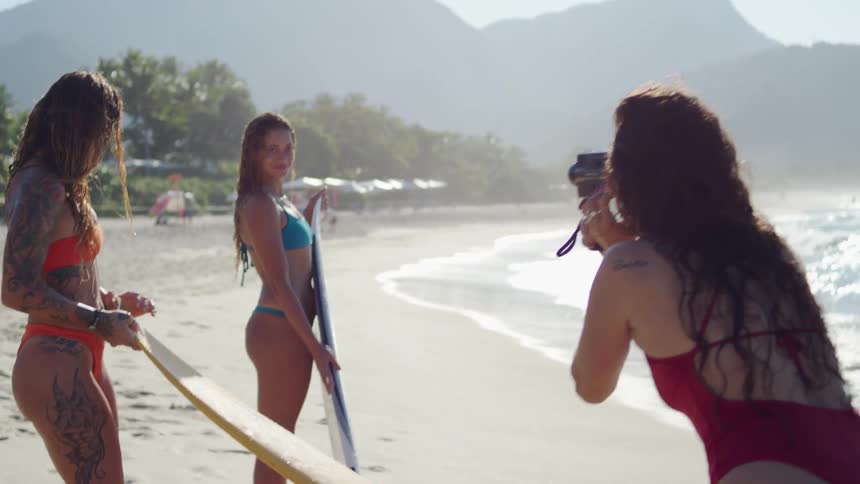 The video shows two women in bikinis standing on a sandy beach next to their surfboards the water is calm with small waves gently rolling in a third person is standing behind them holding a camera