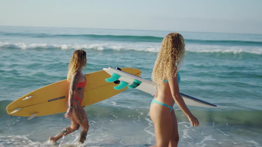 A two young women in bikinis carry surfboards while walking along the beach
