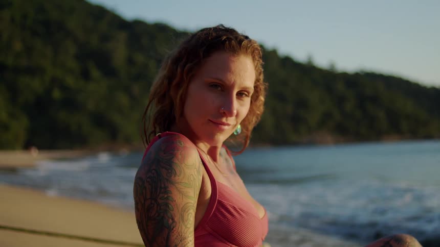 A tattooed woman in a pink bikini sits on the sand by the shoreline with a surfboard beside her illuminated by the golden light of sunset