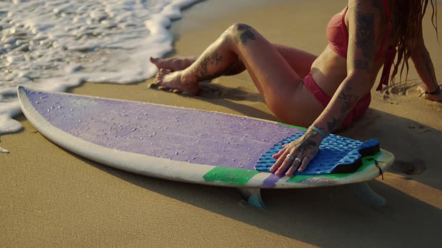 A young woman wearing a bikini sits on the beach with a surfboard by her side as the ocean waves hit her feet and the board at midday