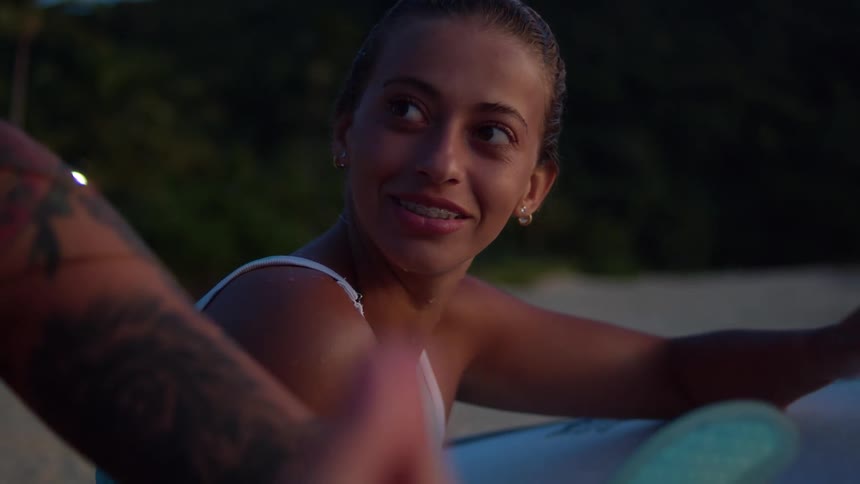 Woman with a surfboard sitting on the sand at the beach talking to another person