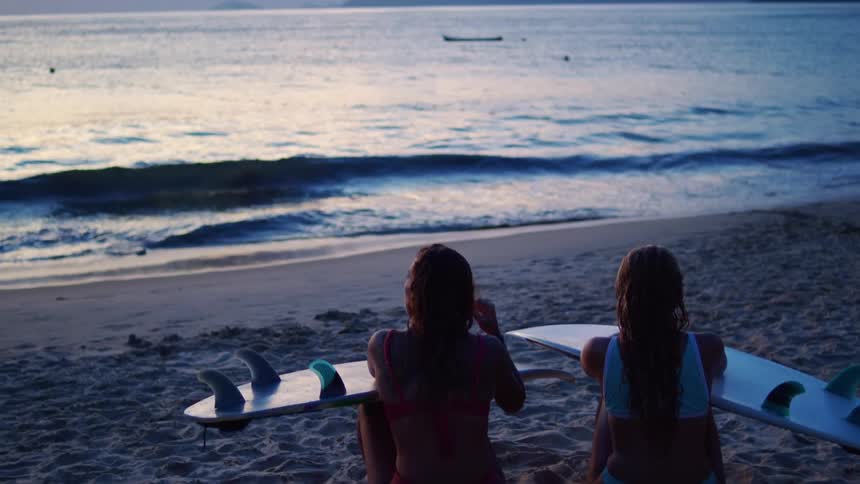 Two women sit on the sand holding surfboards while looking at the sea