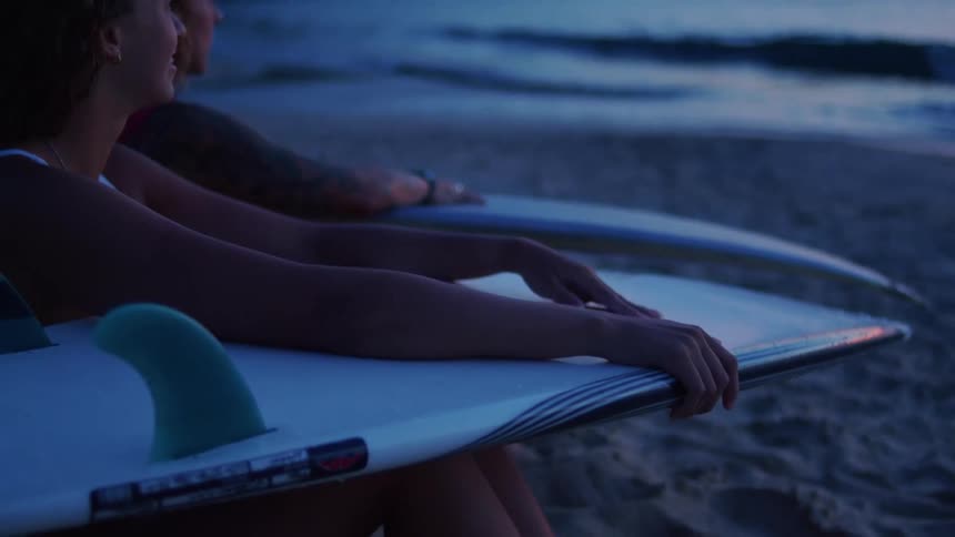 People sitting on the sand hold surfboards at dusk