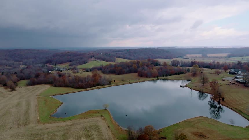 A pond in a rural setting surrounded by grasses and trees with brown leaves from the fall season at midday on a cloudy day