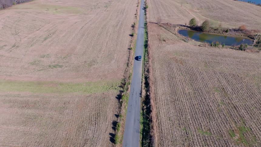 The video shows an aerial view of a car driving down a road surrounded by dry grassy fields there is a small pond or lake to the right of the road