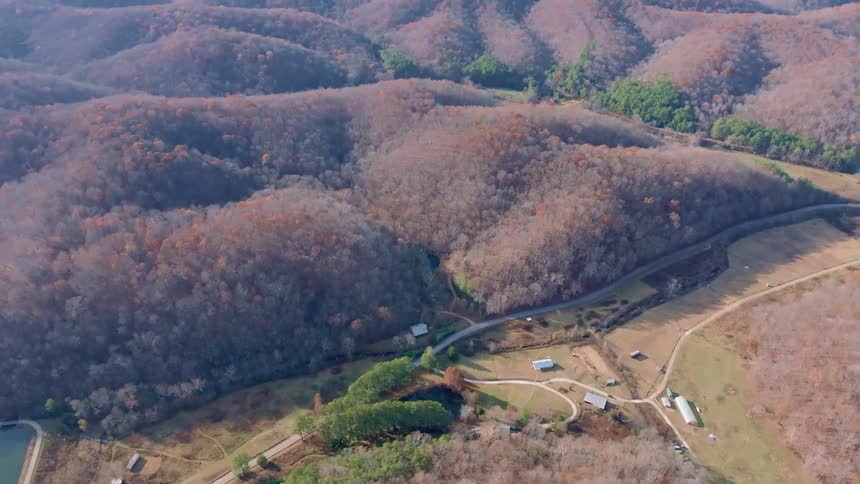 An aerial shot shows a valley surrounded by wooded hills with mostly bare trees a road and a stream wind through the valley floor passing a few buildings and a small pond