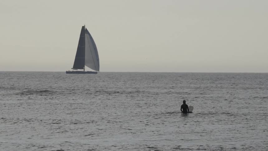 Person with a surfboard in the sea watching a sailboat in the distance