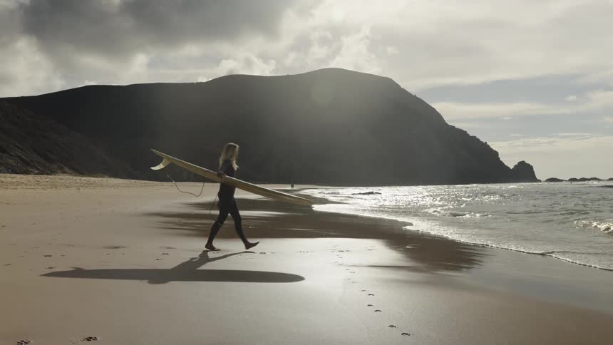 The video shows a person holding a long surfboard and walking toward the ocean on a sandy beach a dog is running in the foreground and there is a large hill in the background the video has a watermark that reads filmsupply