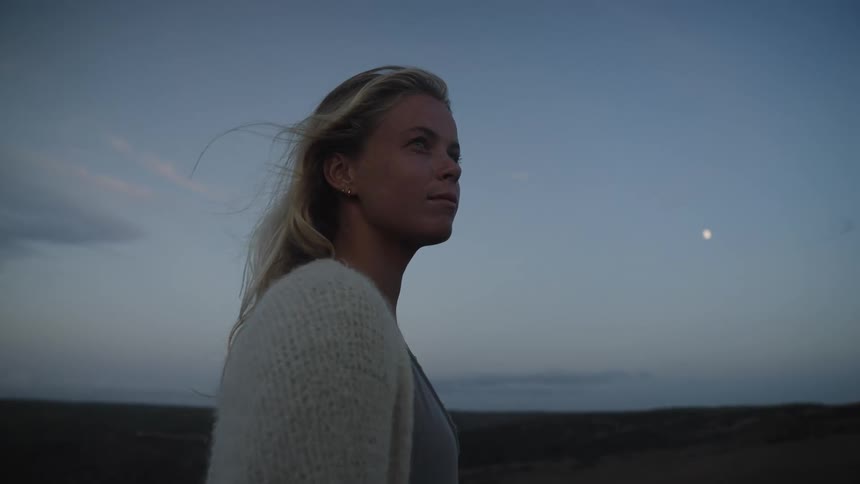 The video shows a side profile shot of a young woman with long blonde hair and a white cardigan standing outdoors at dusk she is looking upwards and her expression is one of calm the background consists of a dark landscape and a bluegray sky with a