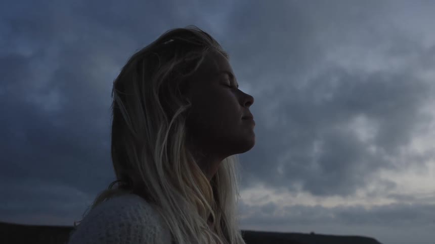 A young woman closes her eyes as a gentle breeze blows through her blonde hair under a clouded sky in the evening