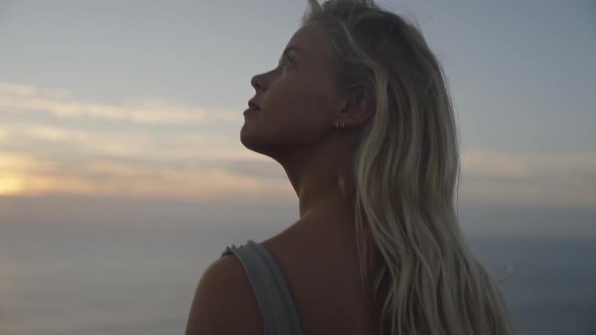 A person with blonde hair is shown from the side with their back to the camera looking out over the ocean at sunset or sunrise they are wearing a lightcolored top and a white knitted shawl draped over their shoulders