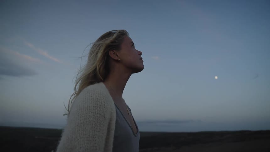 A blonde woman in a light top and cardigan gazes across the landscape at dusk with the moon visible in the sky