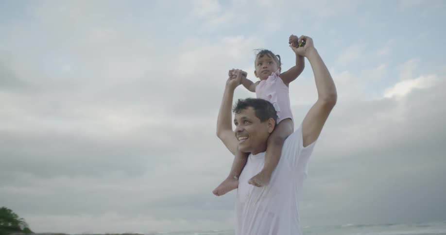 Man carrying toddler girl on shoulders on beach on cloudy day