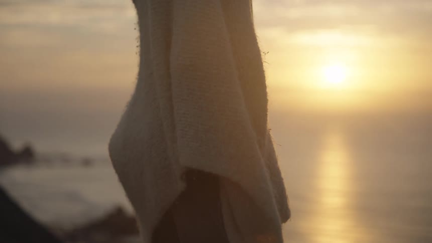 A young woman with blonde hair stands outside at sunset looking out at the ocean she is wearing a lightcolored tank top and a knitted shawl over her shoulders