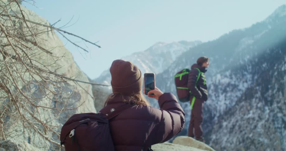 The video shows two people hiking on a snowcovered rocky hill they are both wearing backpacks and one person is taking a picture with a phone