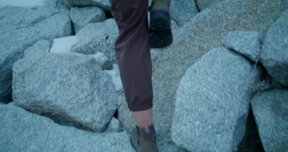 A closeup shot shows a persons feet and legs clad in brown pants and sturdy boots as they carefully walk over large grey rocks there are patches of white snow or ice visible between the rocks suggesting a cold environment