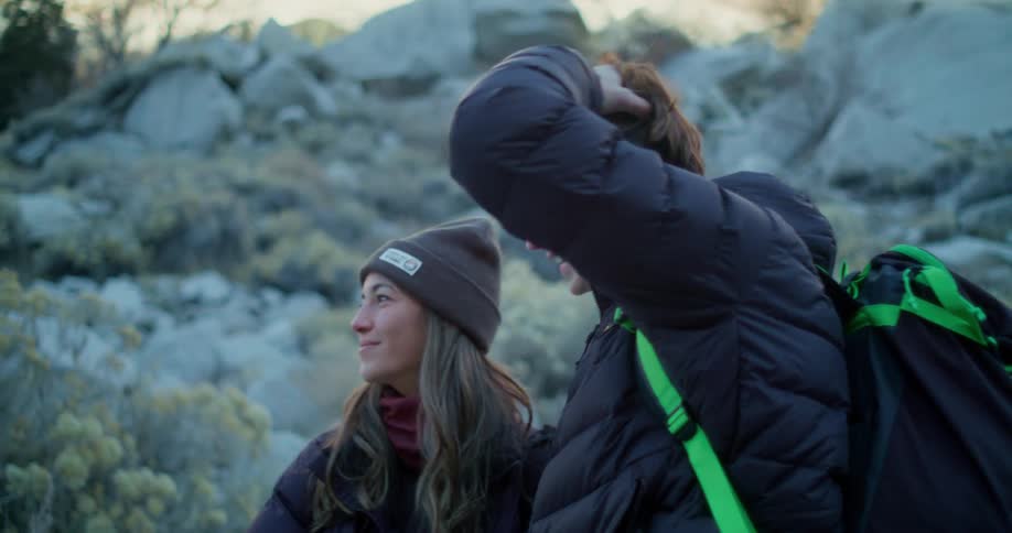 The video shows two hikers a man and a woman standing together in a rocky outdoor landscape they are both wearing dark puffy jackets and are looking offcamera to their right smiling the man has a bright green strap from a backpack visible on his s