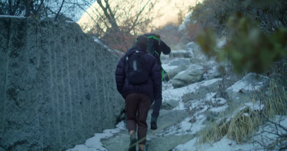 Two women carrying backpacks hike up a mountain trail at midday