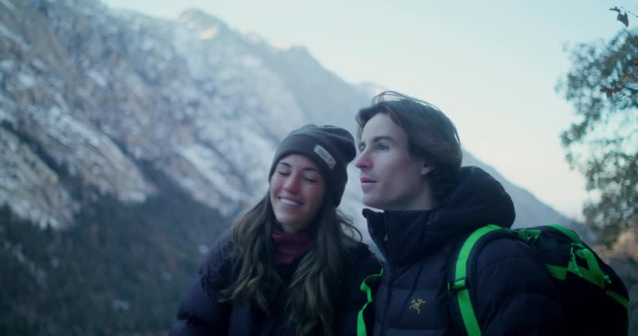 A smiling young couple wearing winter clothes and carrying backpacks looks at a mountain range in front of them and the woman lays her head on his shoulder