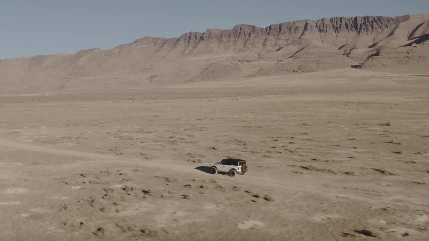 An aerial shot shows a white offroad vehicle driving across a vast flat and arid landscape in the background a large rocky mountain range stretches across the horizon