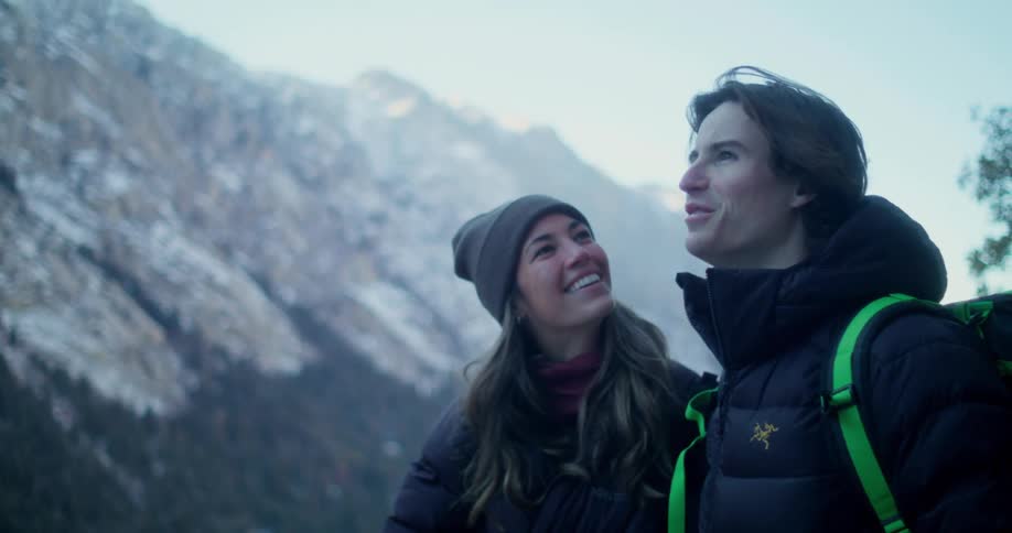 Couple in winter clothing smiles while looking at snowy mountains
