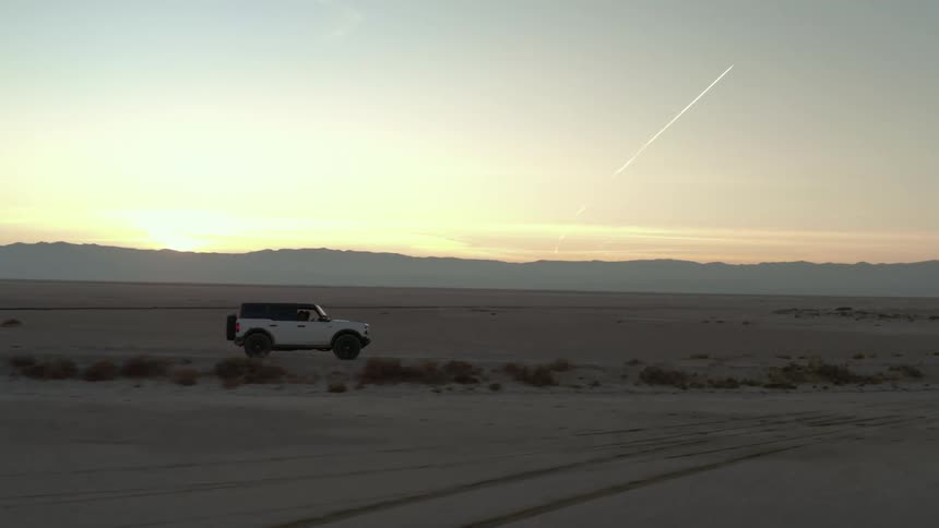 A white suv drives across a barren desert landscape at sunrise with mountains silhouetted in the distance