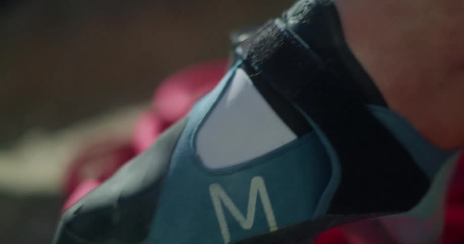 A closeup shows a blue climbing shoe marked with an m with a persons hands putting the shoe on a foot