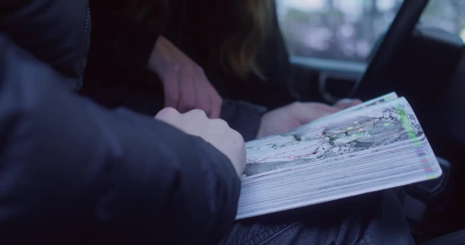 A man and a woman sit behind a car reading both wearing winter jackets at midday