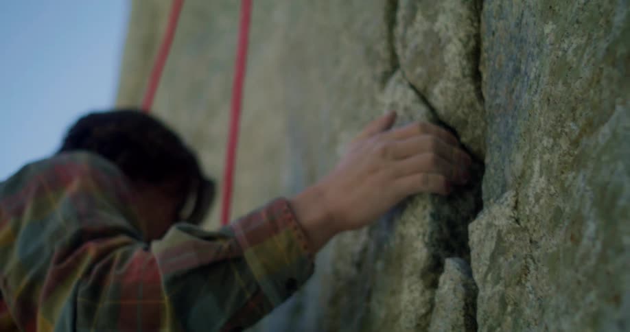 The video shows a lowangle shot of a person rock climbing they are wearing a plaid shirt and their hand is gripping a crevice in a large textured rock face a red rope is visible running vertically alongside them the sky is a clear blue and the water