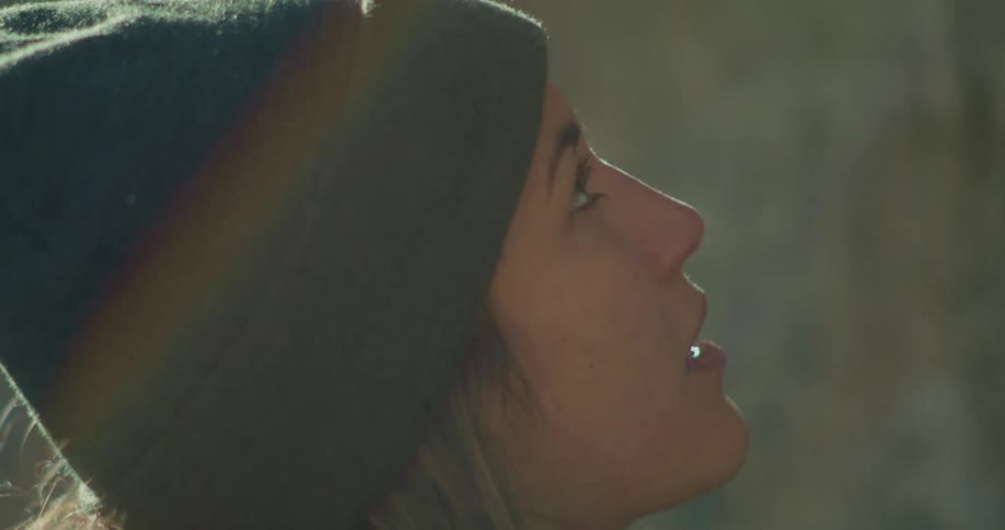 A young woman wearing a head warmer stands outdoors at midday looking up at a rock