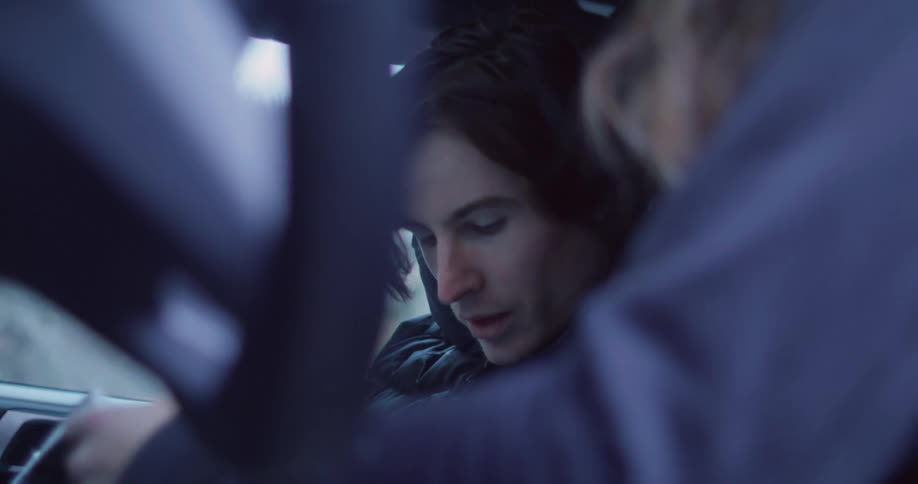 Young man in coat sitting inside a car looking down next to a person