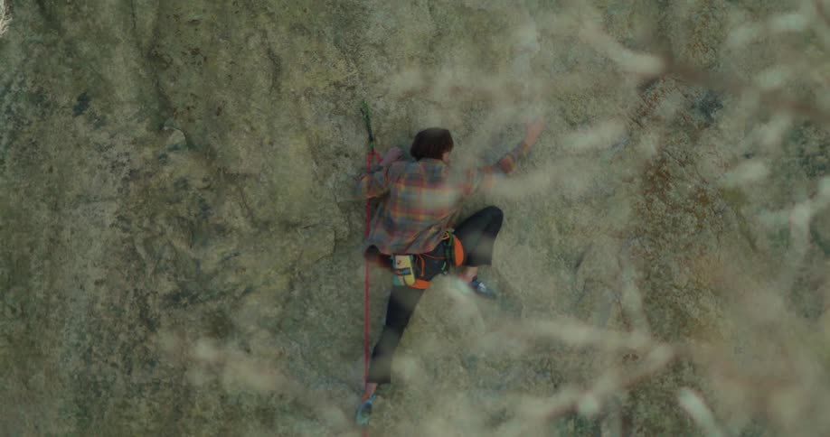 A closeup view from above shows a person in a plaid shirt and climbing harness as they ascend a rock face they are secured by a red rope and are actively reaching for handholds to continue their climb